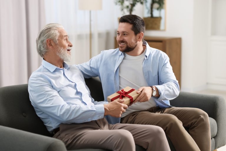 Filho entregando presente ao pai no Dia dos Pais, sentados em um sofá e sorrindo com afeto.