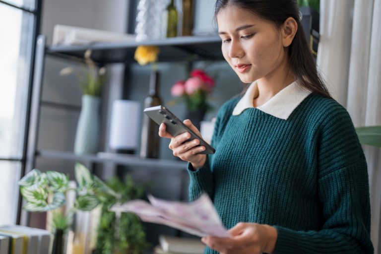 Mulher jovem analisando boletos e usando o celular para organizar suas contas a pagar no ambiente de trabalho.