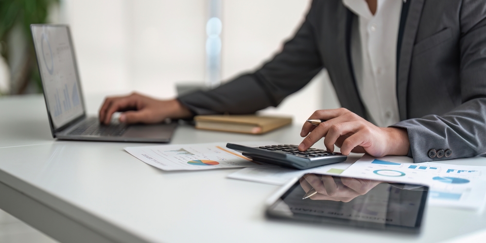 Pessoa utilizando calculadora e computador para analisar gráficos financeiros, representando o processo de como calcular preço de venda de produtos e serviços.