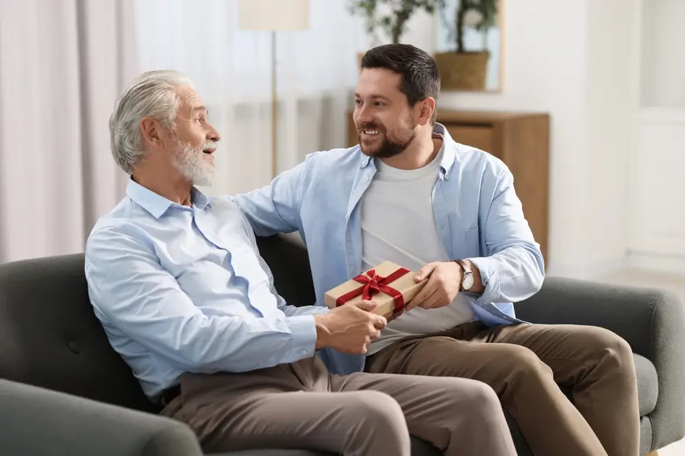 Filho entregando presente ao pai no Dia dos Pais, sentados em um sofá e sorrindo com afeto.