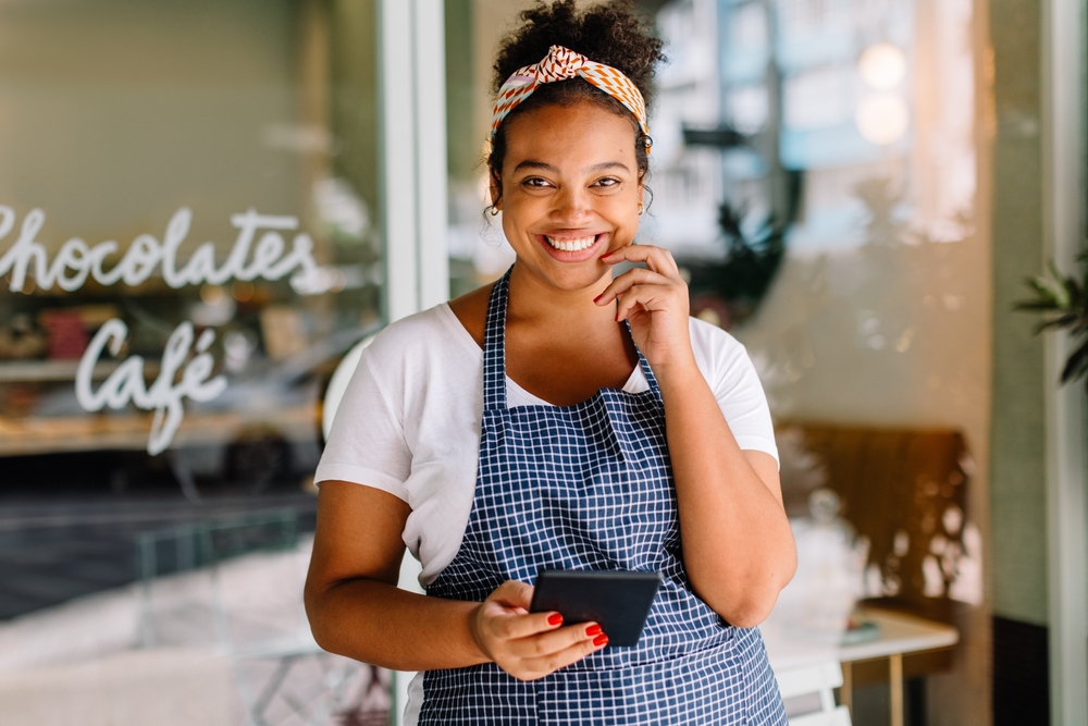 Empreendedora sorridente em frente ao seu café usando o celular para gerenciar o negócio — conceito de gestão de pequenas empresas.