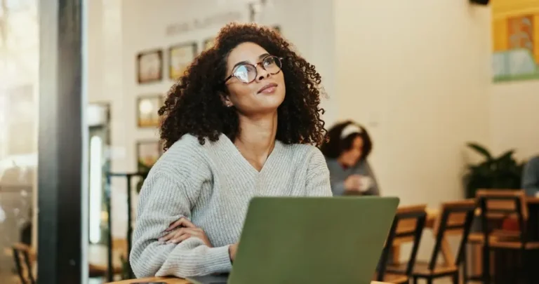 Mulher jovem de óculos, com cabelo cacheado, usando suéter cinza, está sentada em uma cafeteria olhando pensativa para cima, enquanto trabalha em seu notebook.