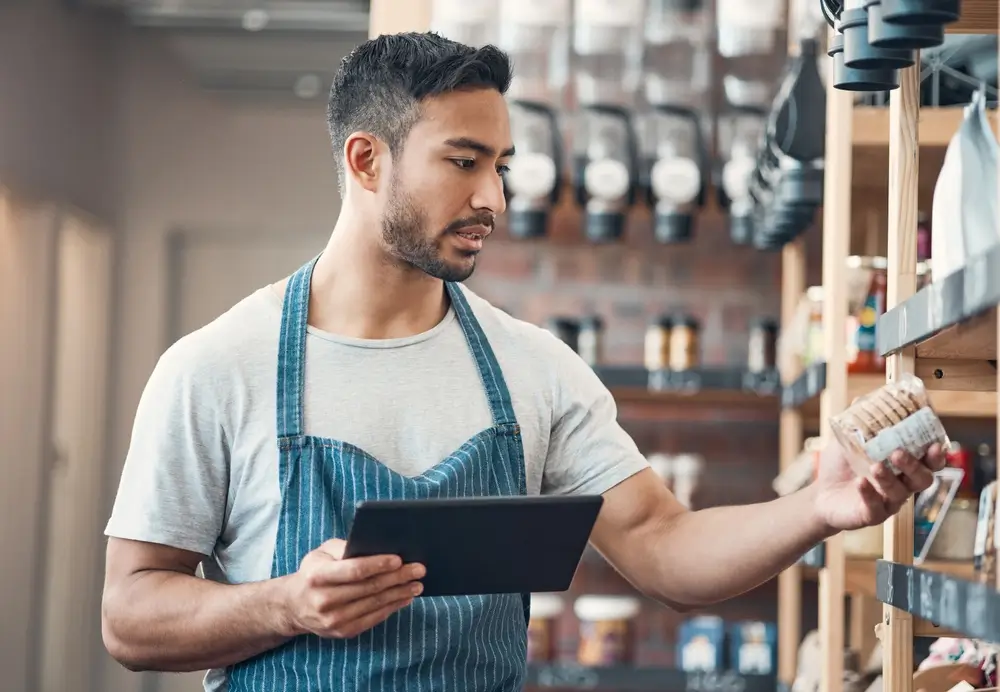 Homem com avental azul segurando um tablet e analisando produtos em prateleira, representando atividades de gestão de estoque.
