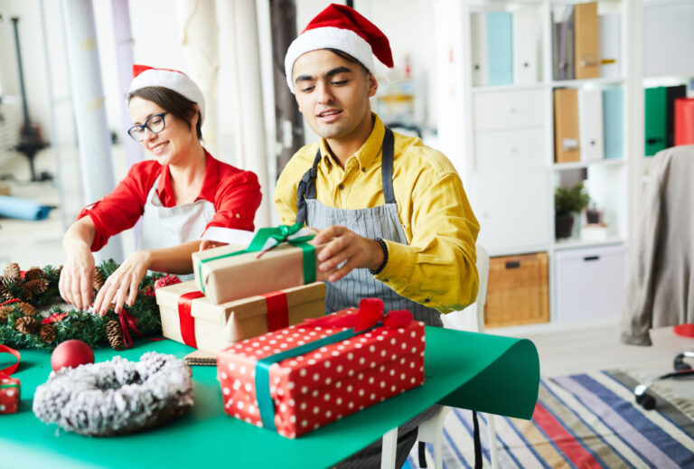 Dois empreendedores com gorros de Natal organizando presentes e decoração enquanto trabalham no planejamento de vendas para o fim do ano.