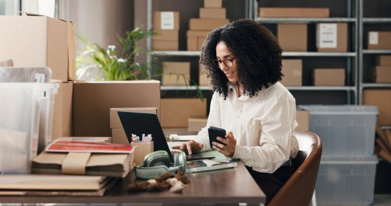 Empreendedora organizando pedidos no notebook e celular em estoque com caixas, representando como centralizar estoque, vendas e finanças em um só lugar.