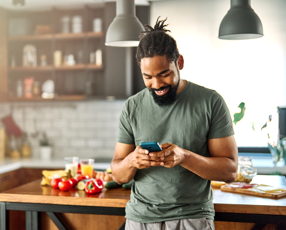 Homem sorrindo mexendo no celular dentro de casa com ingredientes frescos na bancada da cozinha ao fundo, representando negócios para fazer em casa