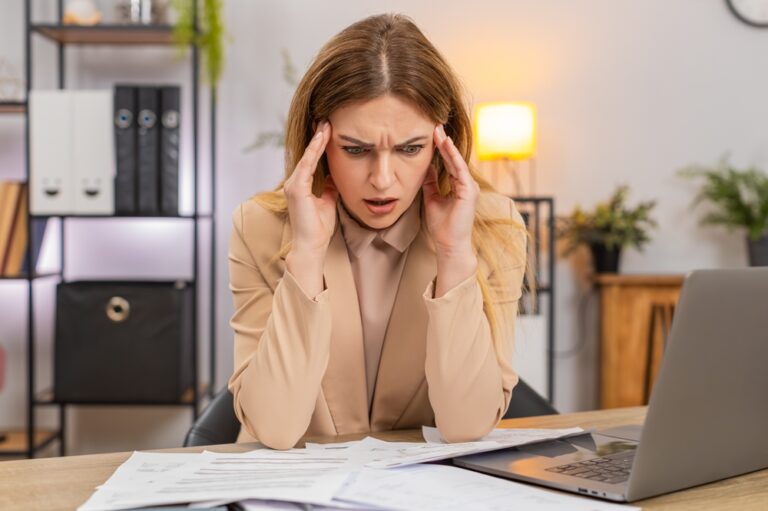 Mulher empreendedora com expressão de estresse diante de papéis e notebook, ilustrando a dificuldade de fazer controle de estoque e vendas sem um app